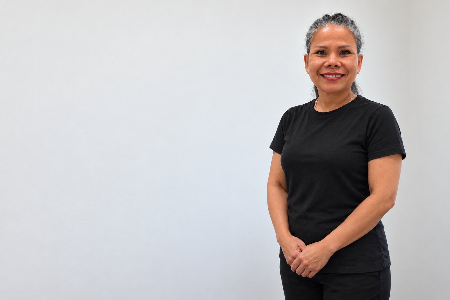 A Thai massage therapist standing against a plain pale ground, arms folded, smiling.